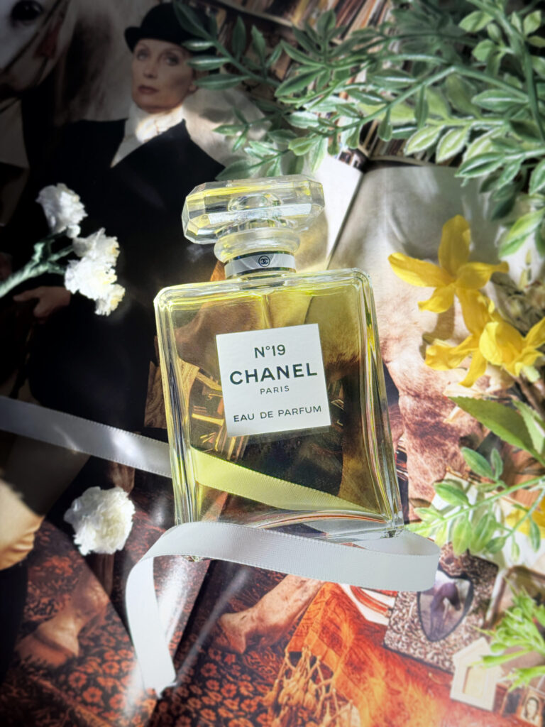 A bottle of Chanel Number 19 wrapped in white ribbon with a variety of flowers next it it. The background is an open book with the image of a woman in riding gear holding the lead of a horse.
