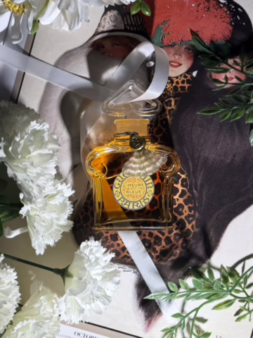 A bottle of Guerlain's L'Heure Bleue wrapped in white ribbon, surrounded by flowers, with a vintage magazine cover in the background.