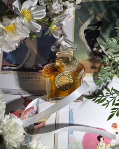 A bottle of Guerlain's L'Heure Bleue wrapped in white ribbon with flowers surrounding it and 1910s Vogue covers in the background.